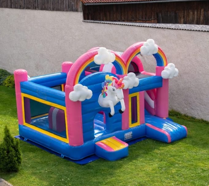Bunte Hüpfburg mit Regenbogen, Wolken und einem Einhorn in einem Garten.